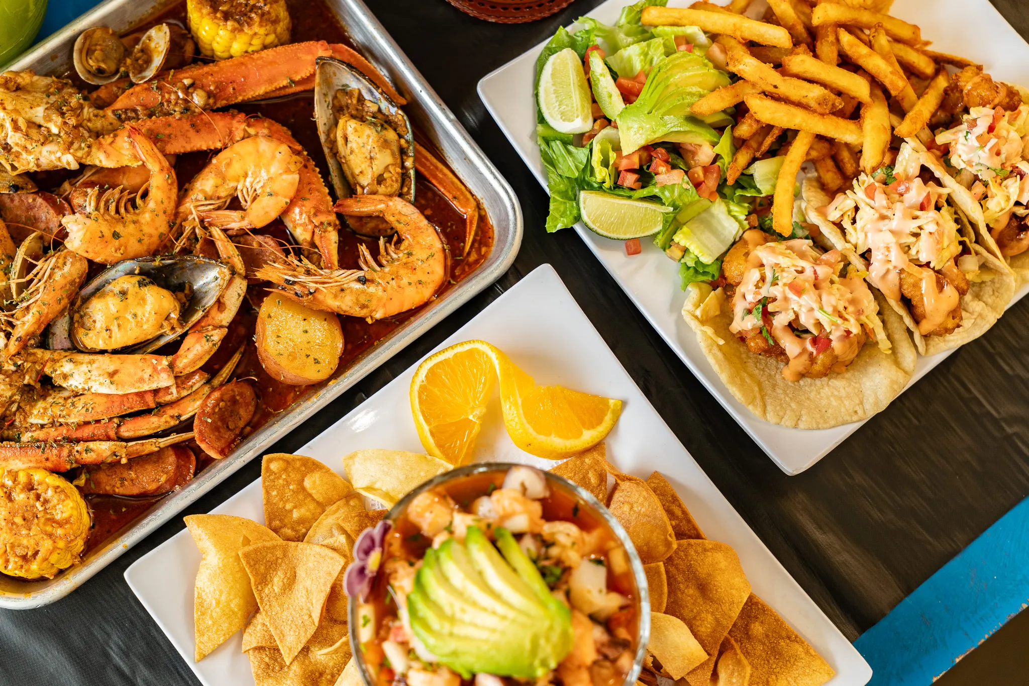 A shareable seafood boil tray at Wacky Crab Shack with shrimp, mussels, crab legs, corn, and potatoes in Cajun sauce, beside shrimp tacos and fries
